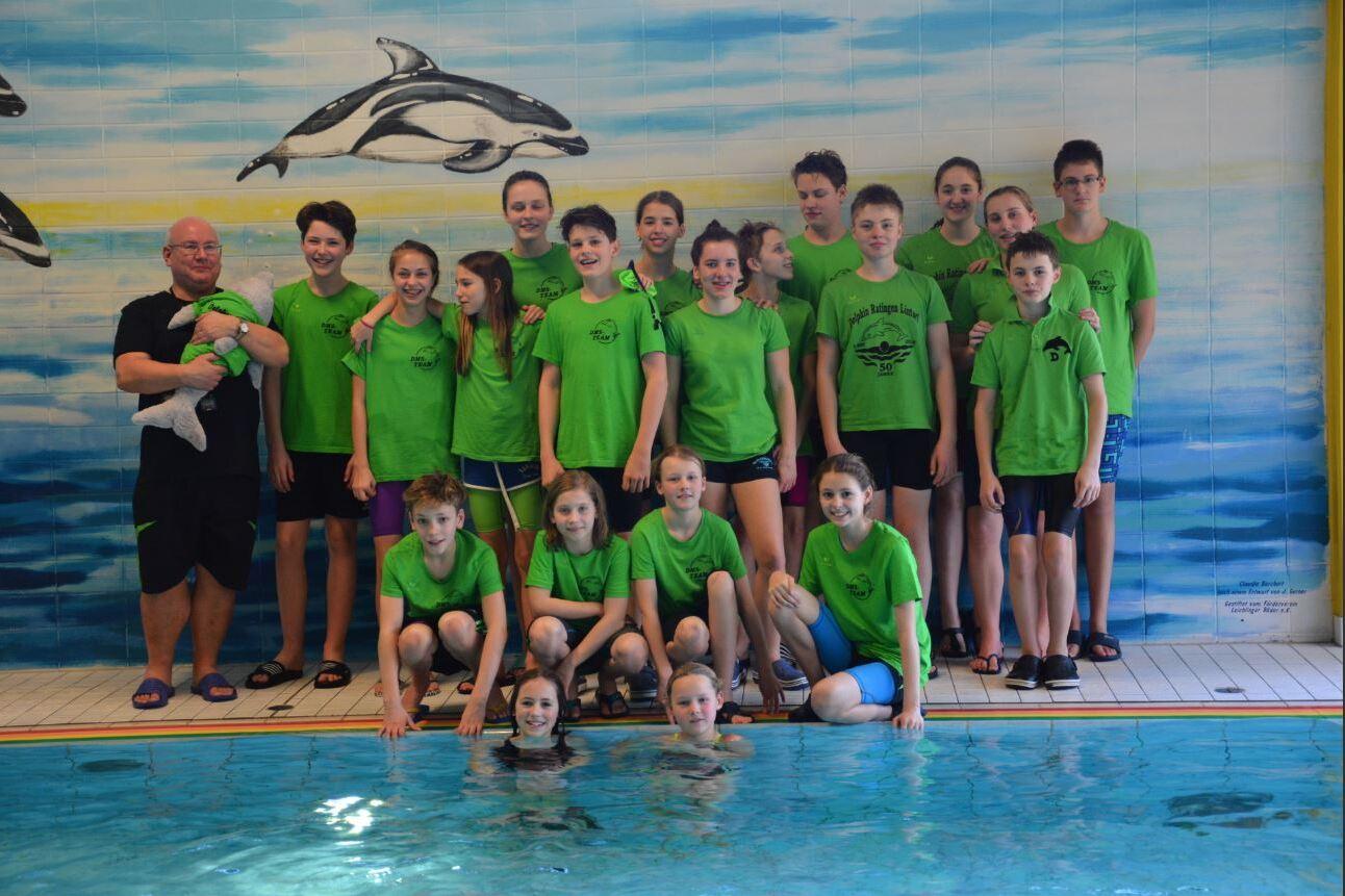 Gruppennfoto von Schwimmern in grünen T-Shirts neben einem Pool, mit einem Trainer und Delfin-Motiven an der Wand.