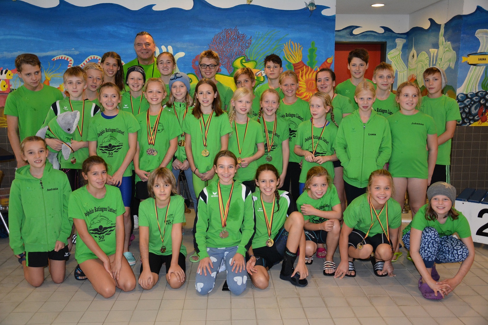 Gruppenfoto von jungen Schwimmern in gr&uuml;nen T-Shirts mit Medaillen, umgeben von einer bunt gestalteten Wand.