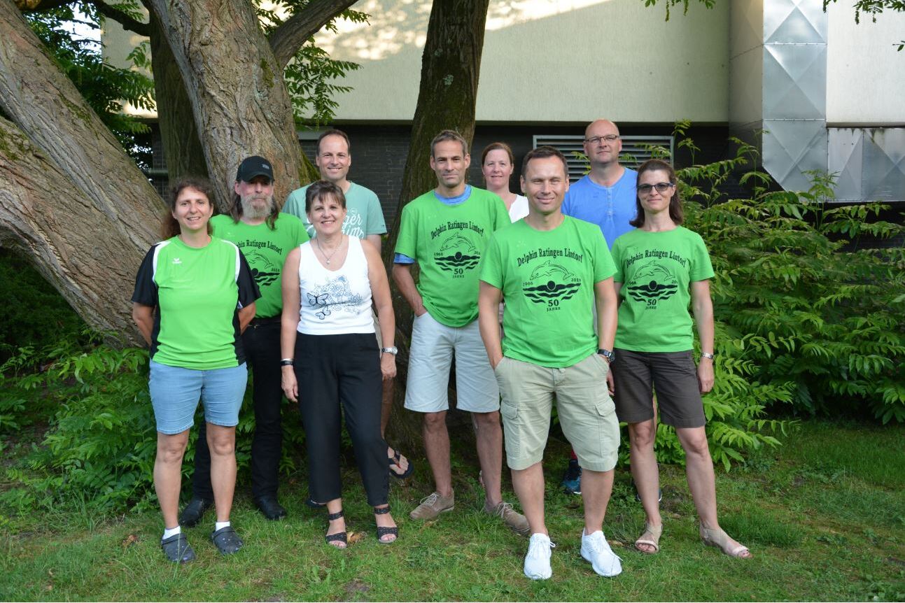 Gruppe von acht Personen in gr&uuml;nen T-Shirts steht unter einem Baum in einer lichtdurchfluteten Umgebung.