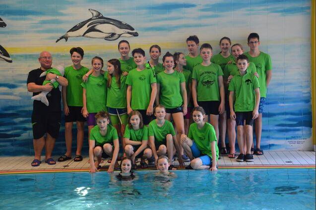 Gruppennfoto von Schwimmern in gr&uuml;nen T-Shirts neben einem Pool, mit einem Trainer und Delfin-Motiven an der Wand.