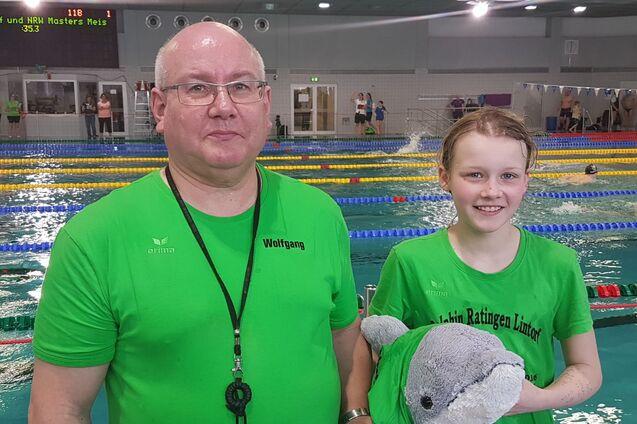 Ein Trainer und ein M&auml;dchen in gr&uuml;nen T-Shirts stehen am Beckenrand mit einem Pl&uuml;schdelfin in der Hand.