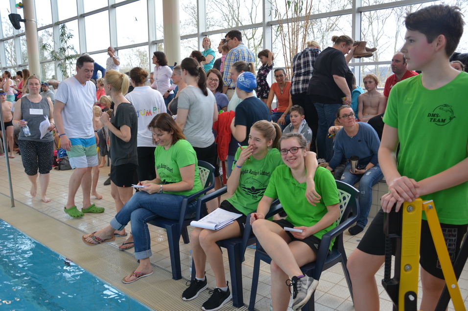 Zusammenkunft von Teilnehmern und Zuschauern bei einem Schwimmwettbewerb, Aufsichtspersonal in gr&uuml;nen Shirts sitzt am Beckenrand.