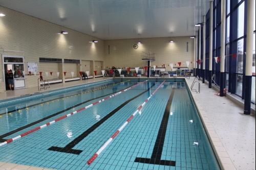 Schwimmhalle mit Sportbecken, drei Bahnen, Schwimmlehrern und Zuschauern am Beckenrand. Gro&szlig;er Fensterbereich f&uuml;r Tageslicht.