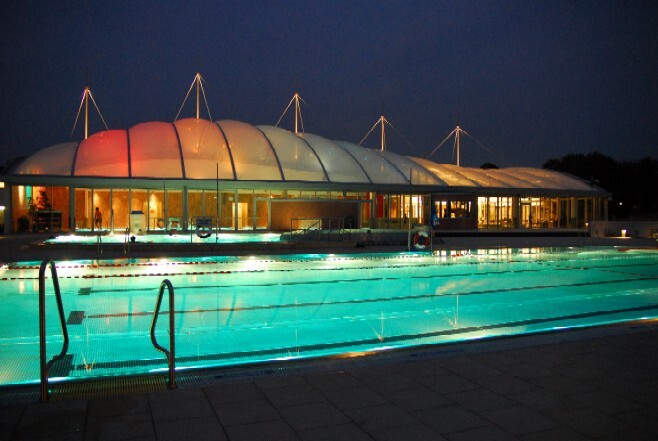 Beleuchtetes Schwimmbad mit gl&auml;sernem Dach und reflektierendem Wasser im Dunkeln.