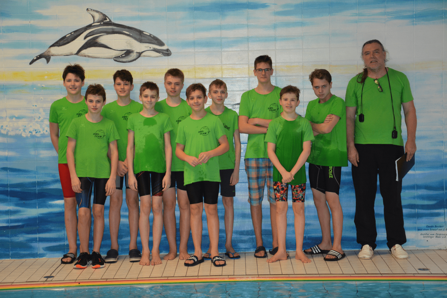 Gruppe von zehn Jugendlichen in gr&uuml;nen T-Shirts, stehend vor einer Wandmalerei von Delfinen in einem Schwimmbad.