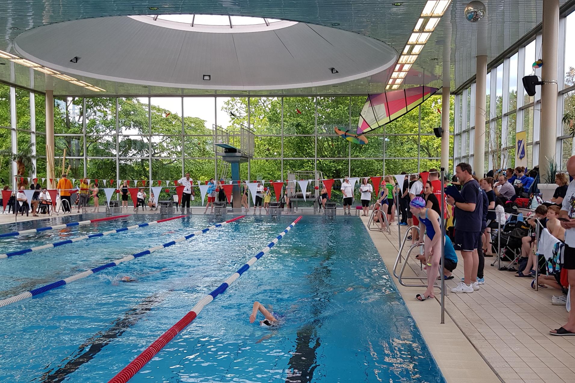 Schwimmbecken in einer Halle mit Wettkämpfen, Zuschauern und Schwimmern in verschiedenen Bahnen.