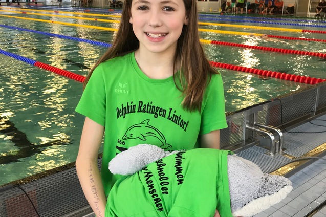 M&auml;dchen in gr&uuml;ner Schwimmausr&uuml;stung h&auml;lt einen Stoffdelfin vor einer Schwimmhalle mit Becken im Hintergrund.