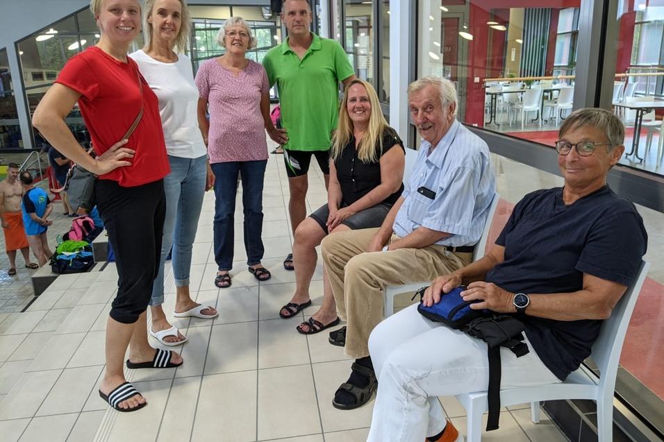 Gruppe von sieben Personen, stehend und sitzend, in einem Sportbereich mit Schwimmbad und B&auml;nken.