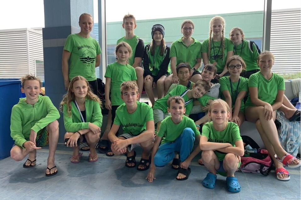 Gruppe junger Schwimmer in gr&uuml;nen T-Shirts sitzt auf einer Bank in einem Schwimmbadbereich.