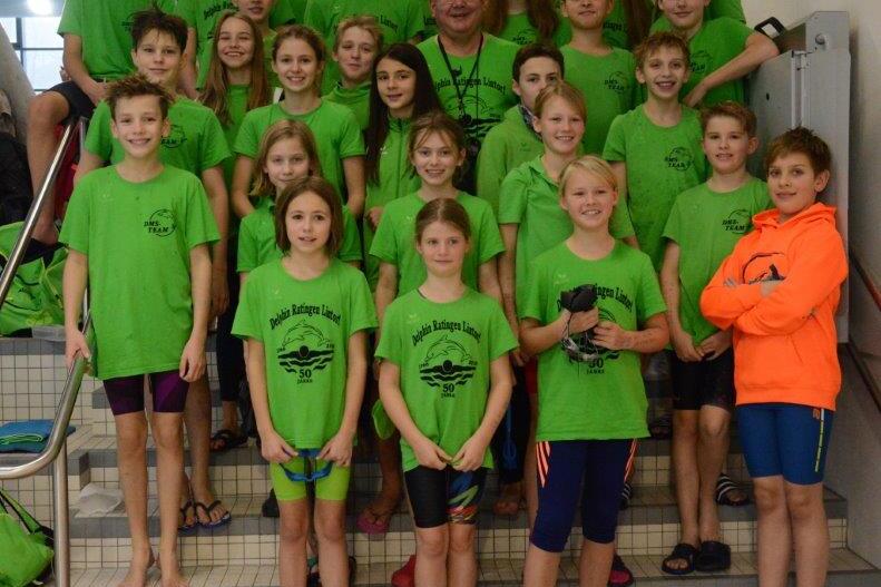 Gruppe von Schwimmern in gr&uuml;nen T-Shirts posiert gemeinsam mit Trainer auf Treppe im Schwimmbad.