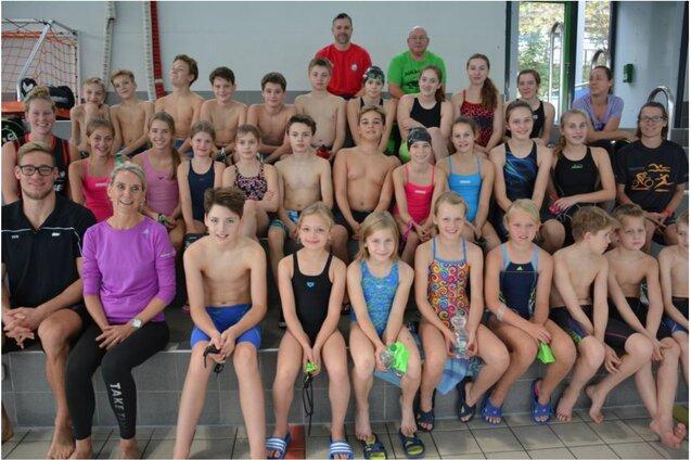 Gruppensfoto von 42 Schwimmern und Trainern, die im Schwimmbadbereich sitzen und l&auml;cheln.