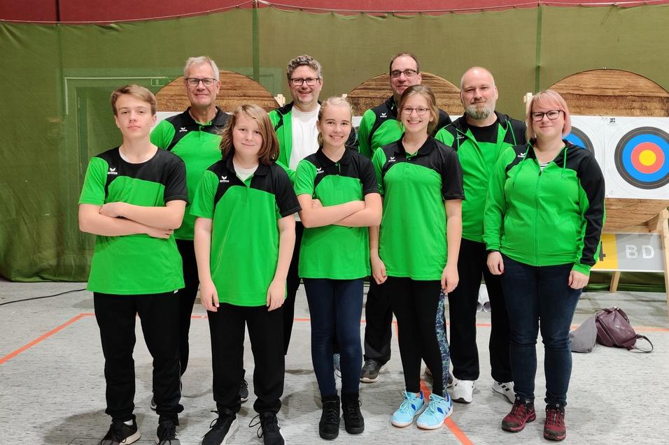 Eine Gruppe von zehn Personen in gr&uuml;nen Sportjacken posiert vor einer Zielscheibe in einer Sporthalle.