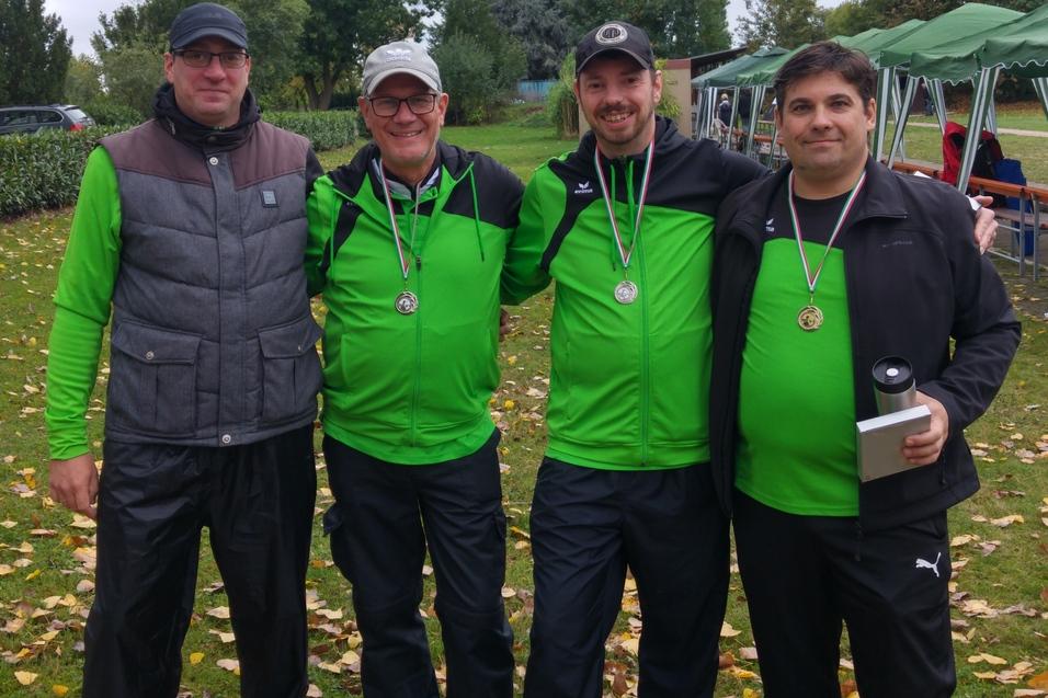 Vier M&auml;nner in gr&uuml;ner Sportbekleidung stehen zusammen im Freien, tragen Medaillen und l&auml;cheln in die Kamera.
