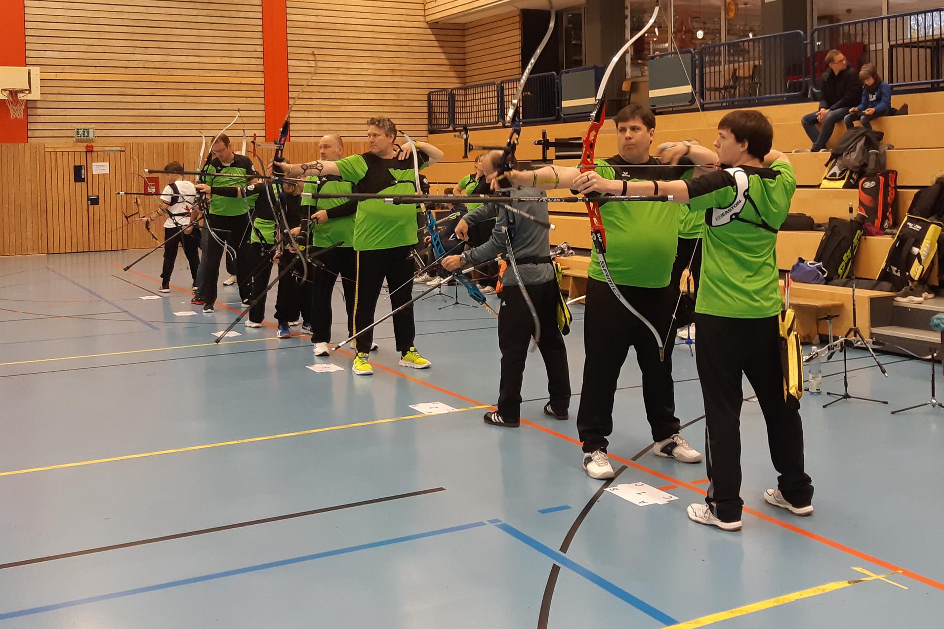 Sechs Bogensch&uuml;tzen in gr&uuml;nen Shirts zielen mit B&ouml;gen auf eine Zielscheibe in einer Halle.