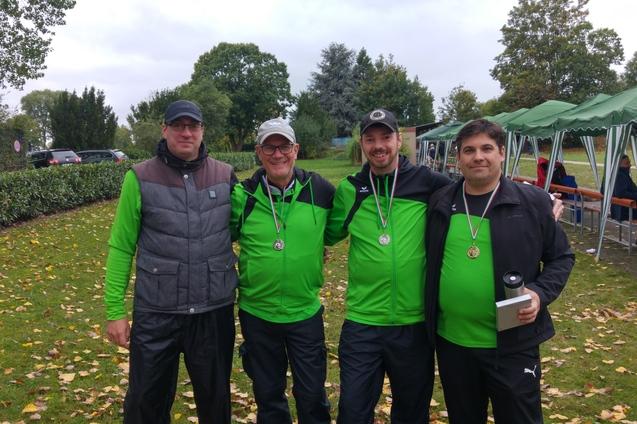 Vier M&auml;nner in gr&uuml;ner Sportbekleidung stehen zusammen im Freien, tragen Medaillen und l&auml;cheln in die Kamera.