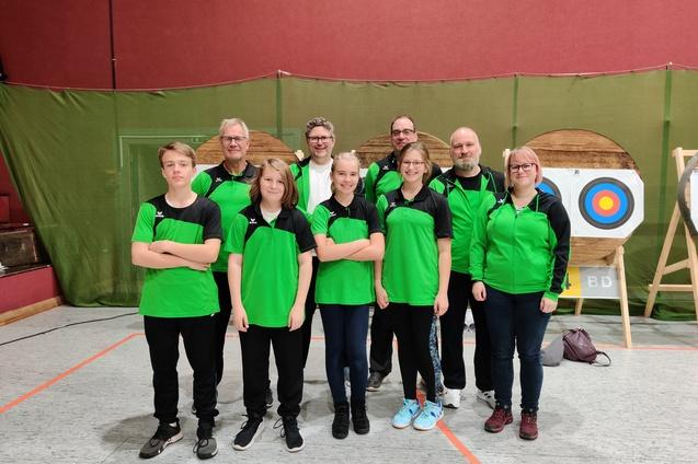 Eine Gruppe von zehn Personen in gr&uuml;nen Sportjacken posiert vor einer Zielscheibe in einer Sporthalle.