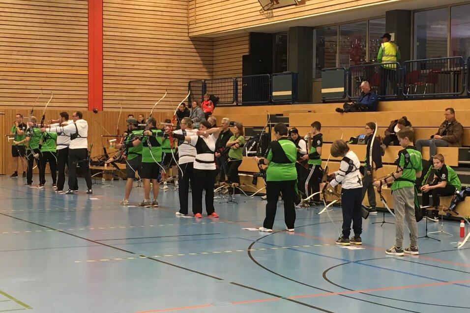 Gruppenschützen mit Bögen in Sportkleidung stehen in einer Halle, Zuschauer sitzen auf Bänken im Hintergrund.