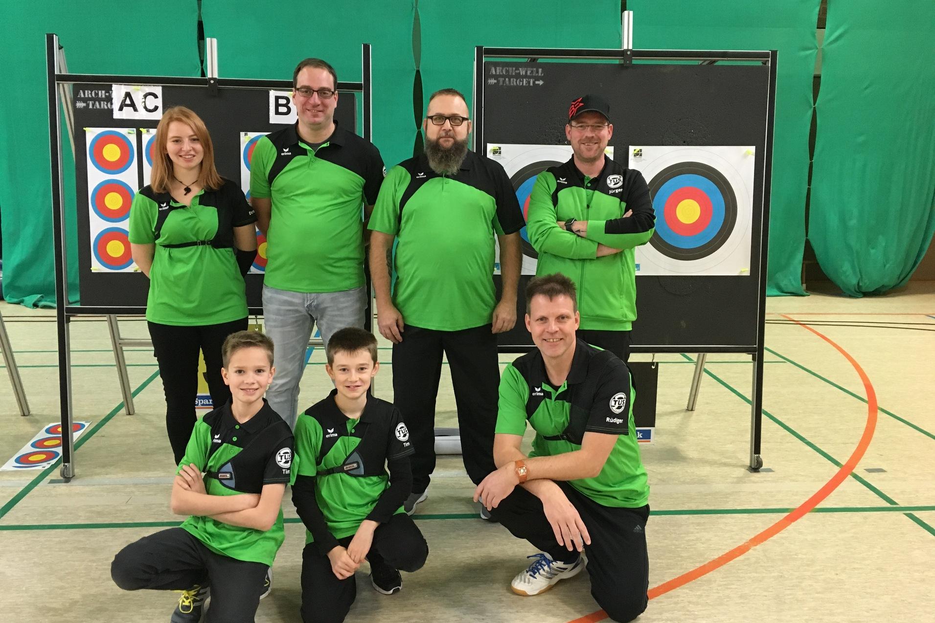 Sechs Personen in gr&uuml;nen Shirts posieren vor Zielscheiben in einer Sporthalle. Zwei Zielscheiben sind sichtbar.