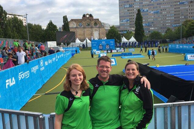 Drei Personen in gr&uuml;nen Sportbekleidungen l&auml;cheln vor einem Wettkampfort mit Zuschauern und Sportanlagen im Hintergrund.