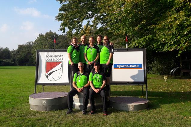 Sechs Personen in gr&uuml;nen Shirts stehen auf einem Podest neben Schildern des Bogensports und der Sparda-Bank.