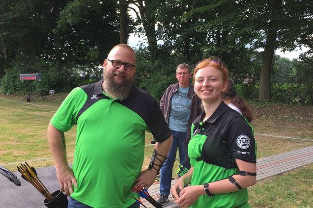 Zwei Bogensch&uuml;tzen in gr&uuml;ner Sportkleidung l&auml;cheln vor einem &Uuml;bungsplatz, ein weiterer Mann steht im Hintergrund.