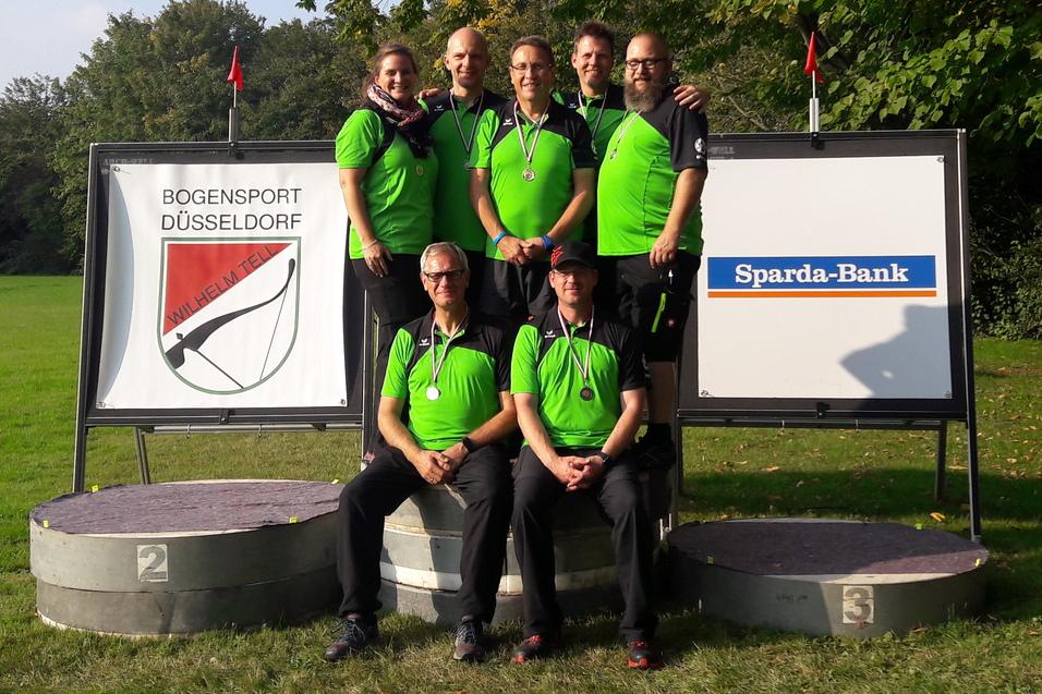 Sechs Personen in gr&uuml;nen Shirts stehen auf einem Podest neben Schildern des Bogensports und der Sparda-Bank.