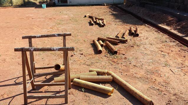 Holzstämme liegen verstreut auf einem trockenen, roten Boden neben einem Holzgerüst. Im Hintergrund ist ein Gebäude sichtbar.