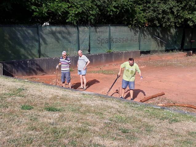Drei Männer stehen auf einem Sportplatz und arbeiten mit Werkzeugen am Boden. Im Hintergrund ist ein Zaun sichtbar.