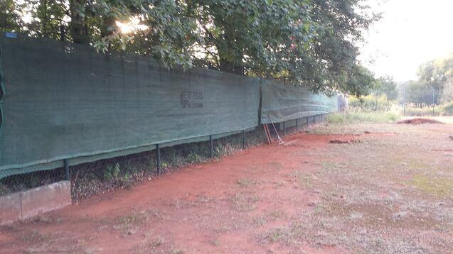 Grüner Zaun entlang eines Tennisplatzes mit erdigem Boden und Schatten von Bäumen im Hintergrund.