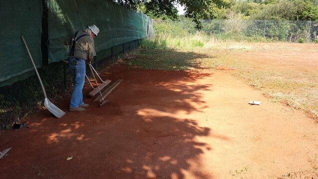 Mann mit Schaufel und Rechen bearbeitet den Boden auf einem ungepflegten Platz, umgeben von Büschen und einem grünen Netz.