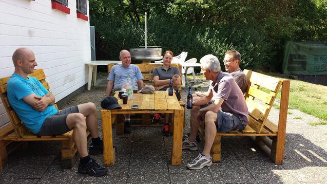Fünf Männer sitzen entspannt auf Holzbänken um einen Tisch im Freien, im Hintergrund grüne Pflanzen.