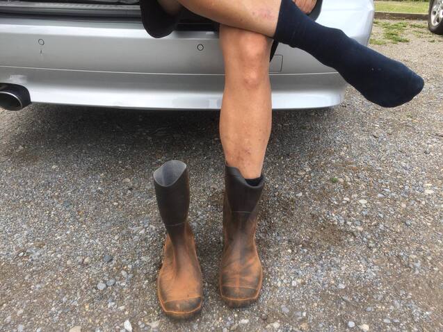 Ein Bein in einem Socken und ein Bein in einem Gummstiefel stehen vor einem Auto auf Kiesboden.