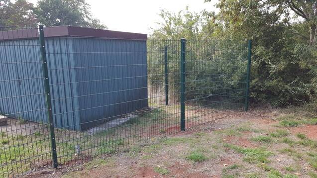 Zaun und grauer Geräteschuppen auf einem unebenem Grundstück mit grüner Vegetation im Hintergrund.
