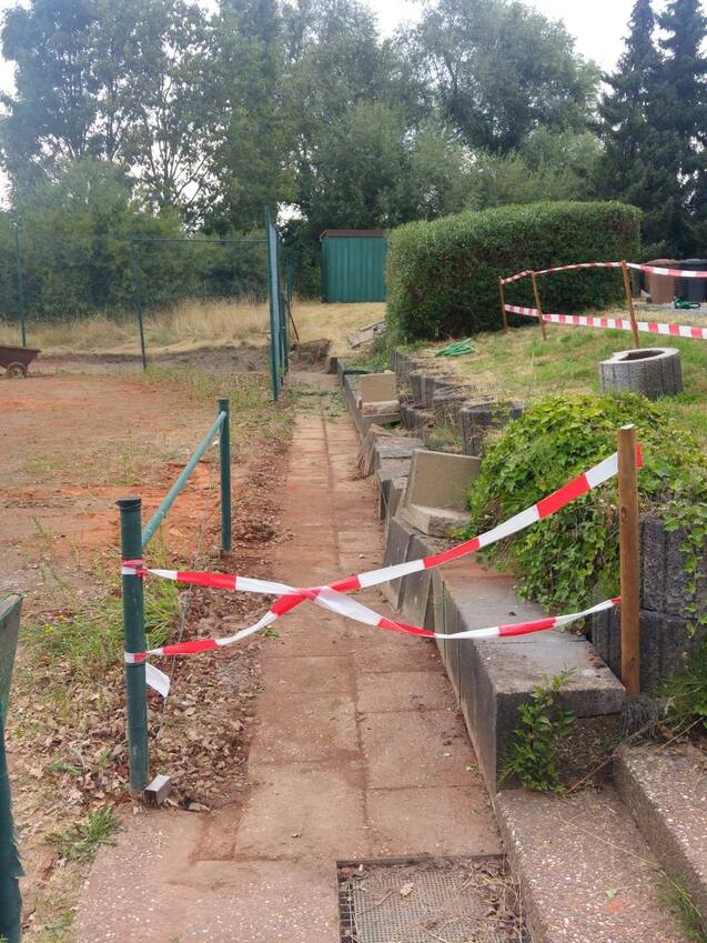 Abgesperrter Weg mit rot-weißer Warnbändchen und Betonstufen, umgeben von Büschen und Bäumen.