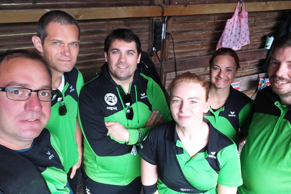 Gruppe von sechs Personen in gr&uuml;nen Sporttrikots posiert gemeinsam in einer Holzunterkunft.