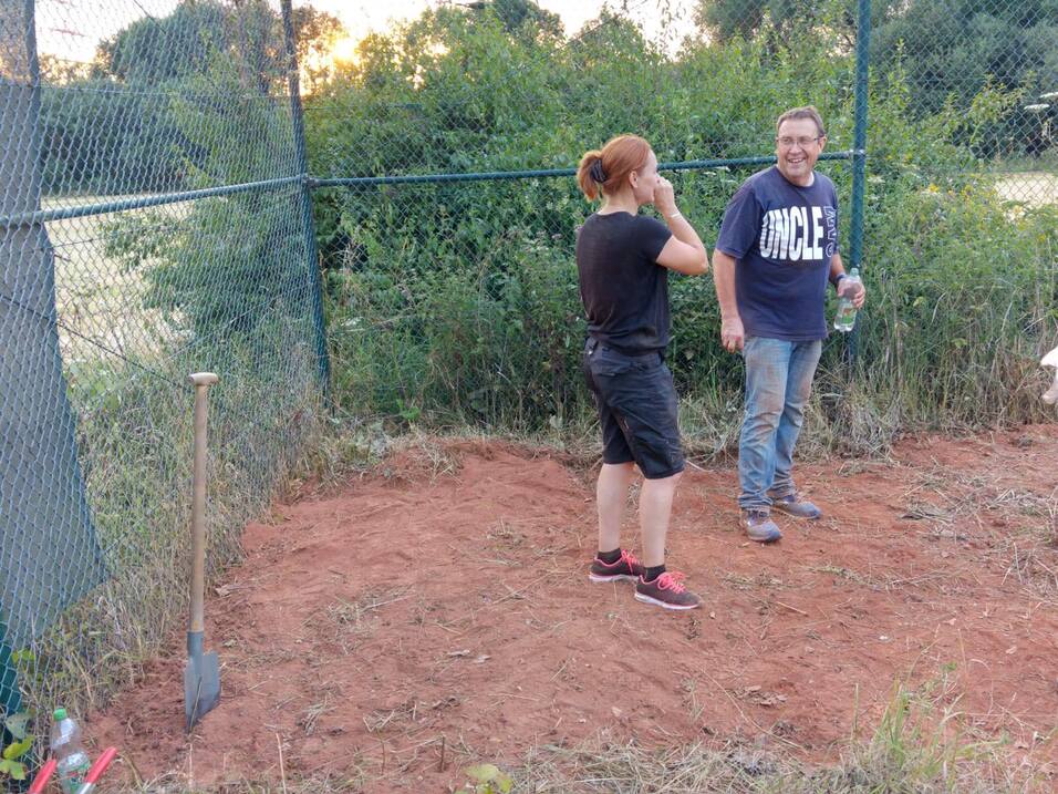 Zwei Personen stehen auf einer unebenen Fläche, umgeben von einem Zaun, eine Schaufel steht im Hintergrund.