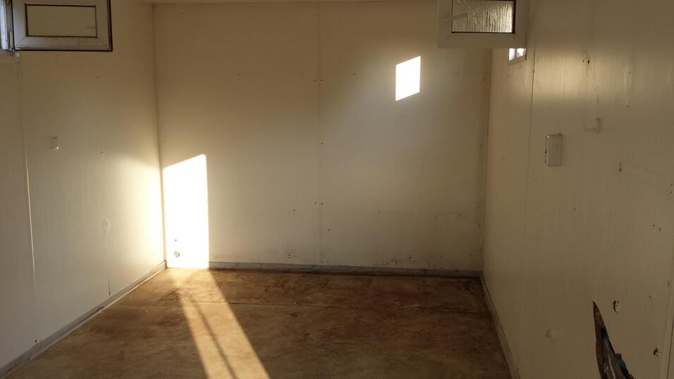 Helle, leere Innenraum mit weißen Wänden, schmalem Fenster und Sonnenlicht, das diagonal auf den Boden fällt.