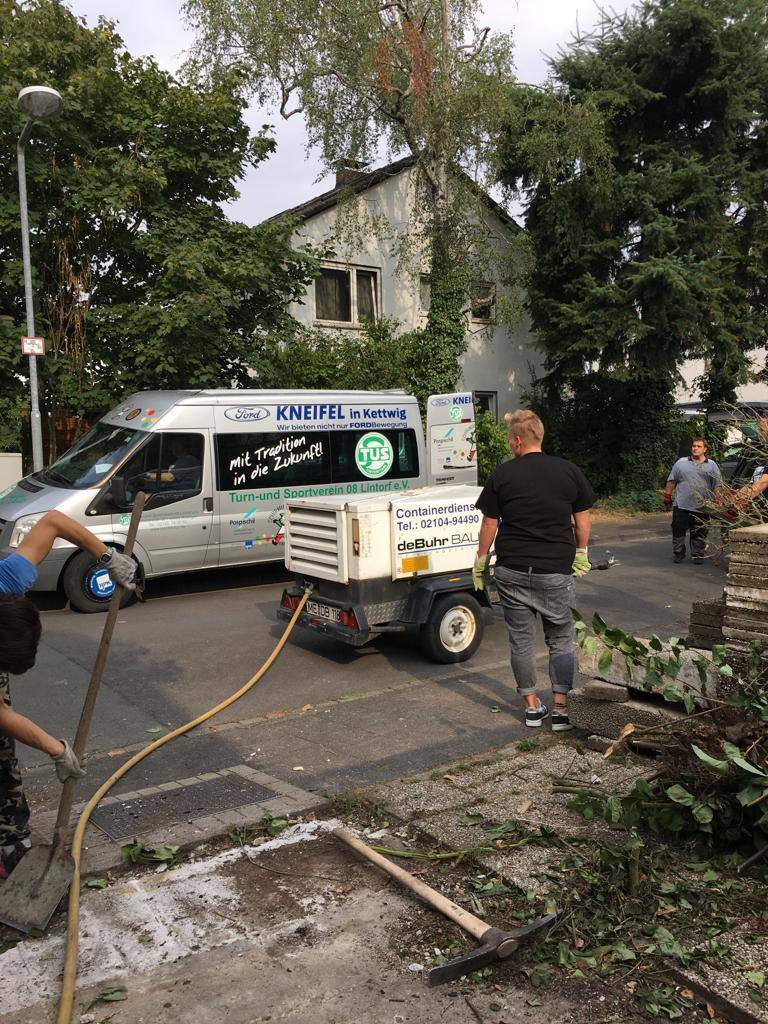 Bauarbeiter räumen Laub mit Schaufeln und Besen, ein Transporter steht vor einem Wohnhaus im Hintergrund.