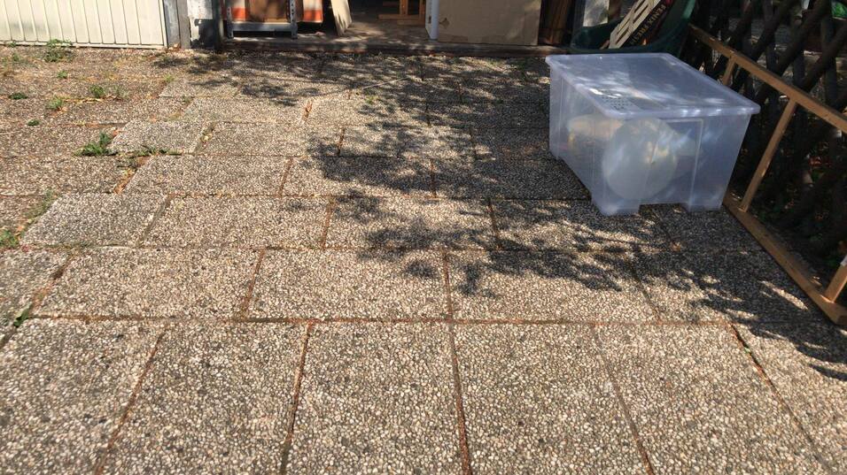 Sicht auf einen grauen, gepflasterten Hof mit einer transparenten Kunststoffbox und Schatten auf dem Boden.