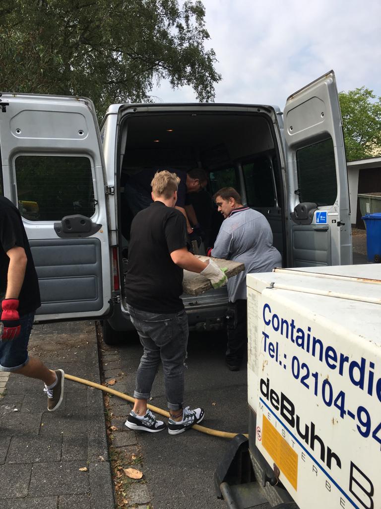 Vier Personen laden Kartons in einen grauen Lieferwagen an einer Straße, während ein Container im Vordergrund steht.