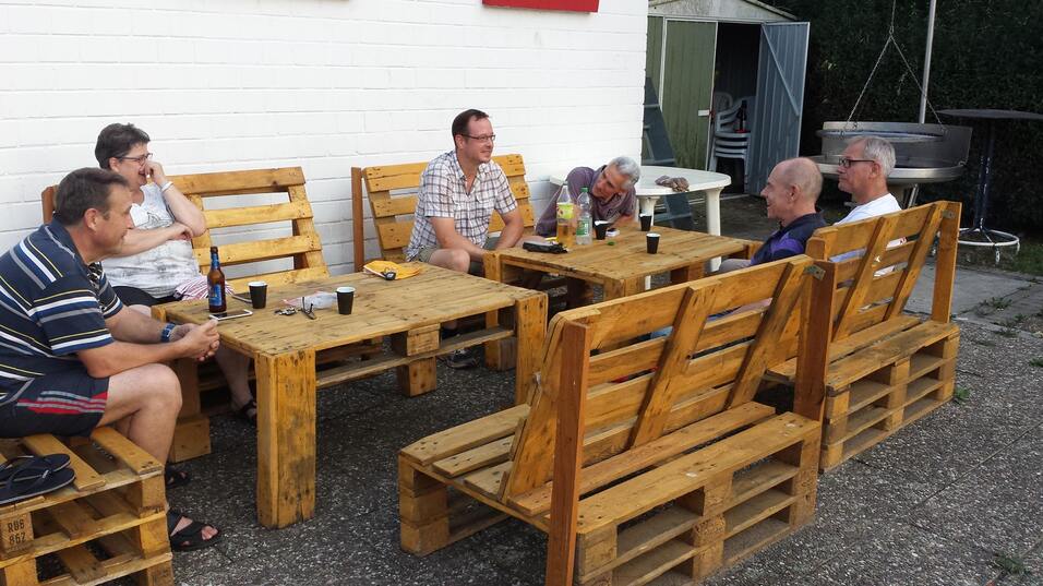 Sechs Männer sitzen an einem Palettentisch und unterhalten sich, umgeben von einfachen Palettenbänken.