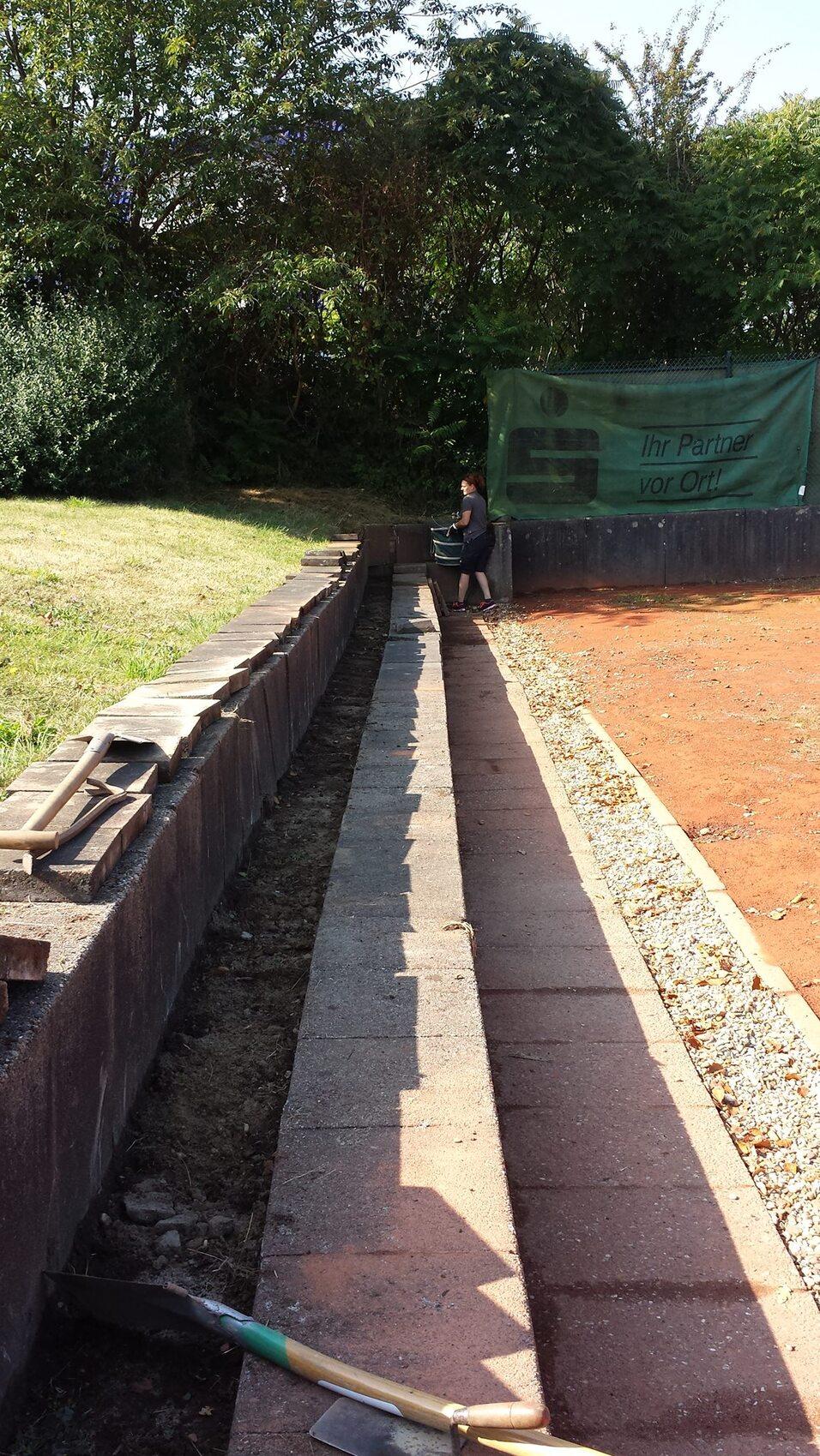 Blick auf eine lange, schmale Baugrube mit Betonsteinen und einer Person, die neben einem stabilen Zaun sitzt.