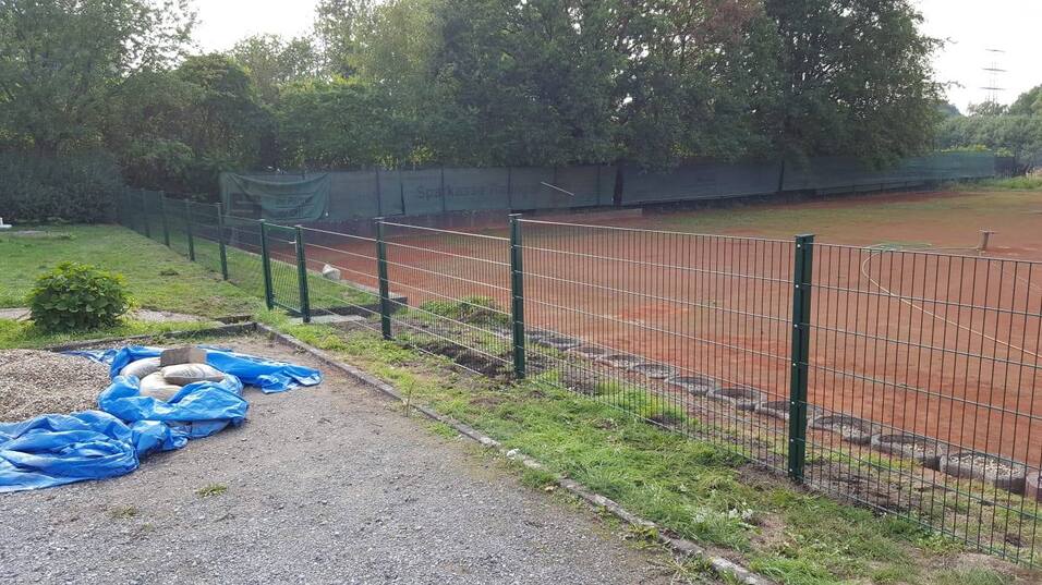 Blick auf einen Tennisplatz mit roter Erde, umgeben von einem Zaun und Bäumen im Hintergrund.