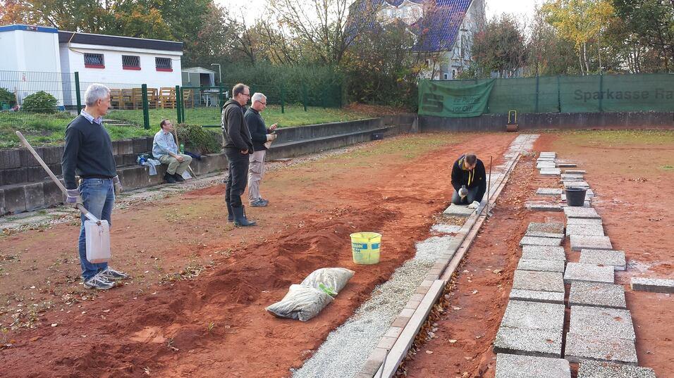 Fünf Personen arbeiten auf einem Tennisplatz, während eine Person Pflastersteine verlegt und andere zuschauen.