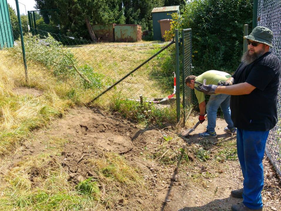 Zwei Männer arbeiten an einem Zaun, einer hält eine Gartenschaufel, der andere mit einer Motorsense auf einem unebenen Boden.