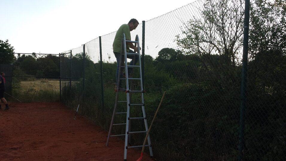 Mann auf einer Leiter repariert einen Zaun an einem Tennisplatz, umgeben von grüner Vegetation und rotem Sandboden.