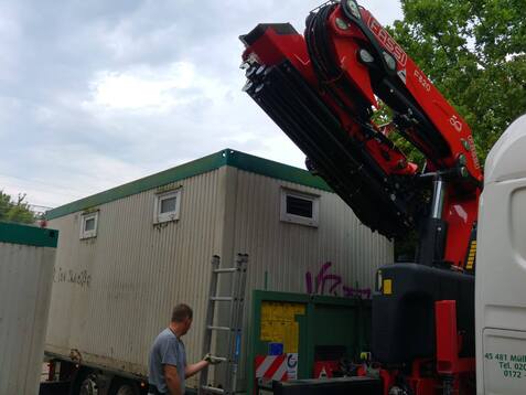 Arbeiter steuert einen Kran, der eine Baucontainer auf einen Lkw hebt, während eine Leiter daneben steht.