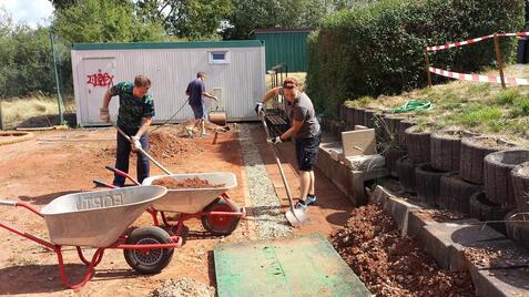 Vier Personen arbeiten im Freien und transportieren Erde mit Schubkarren auf einer Baustelle. Hintergründe sind teilweise eingezäunt.