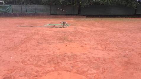 Leerer Tennisplatz mit rotem Sandboden und einem Bewässerungsschlauch in der Mitte.