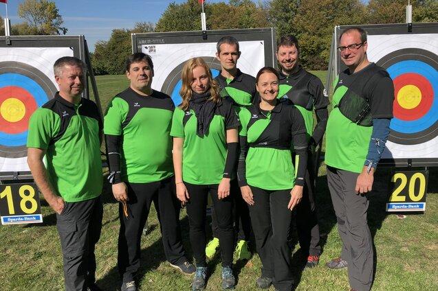 Gruppe von sieben Bogensch&uuml;tzen in gr&uuml;nen Shirts vor zwei Zielscheiben auf einem Rasenplatz.